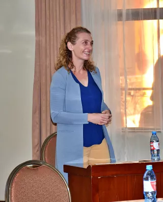 A woman with curly hair stands smiling beside a wooden table in a room with beige curtains.