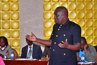 A man in a black buttoned coat stands addressing a group during a meeting. The background features a textured yellow wall