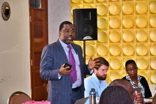 A man in a suit speaks confidently to a seated audience. He holds a phone in his hand.