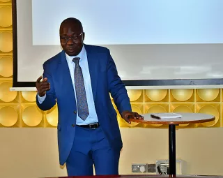 A man in a blue suit gestures while presenting, standing by a round table with a notepad. 