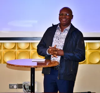Smiling man in glasses, dark jacket, and patterned shirt, stands by a round wooden table with a notepad, in front of a bright projector screen.