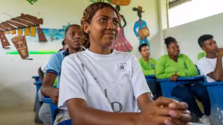 A young girl sitting at a desk in a classroom, smiling among peers.