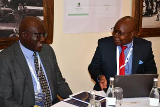 Two men in suits sit at a table, smiling, engaged in conversation.