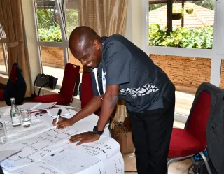 A man stands at a table covered with papers, writing on a large piece of paper.