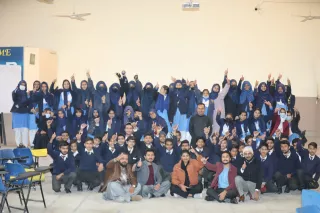 A large group of students and teachers in blue uniforms pose indoors, smiling and raising their fingers in celebration, conveying unity and happiness.