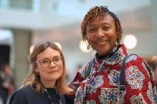 Two women smiling warmly, standing together in a brightly lit indoor setting. One wears glasses and a dark top; the other, a colourful patterned shirt.
