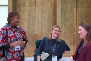 Three people in a lively conversation, smiling warmly. They wear conference badges, standing against a wooden backdrop, conveying a positive, engaging tone.