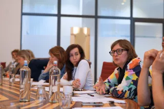 A group of people in a conference room discuss around a table. Papers, pens, and water bottles are visible. The atmosphere appears engaged and focused.