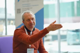 A man with a beard, wearing a brown sweater, gestures while speaking. He's seated indoors at an event, conveying a thoughtful expression.
