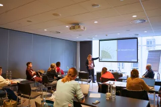 A woman presents at a conference room, pointing to a screen displaying "De Zonnestralen." Attendees sit attentively, creating a focused atmosphere.