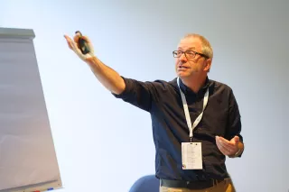 A man wearing glasses and a dark shirt speaks energetically while gesturing toward a flip chart in a bright room. He appears engaged in a presentation.