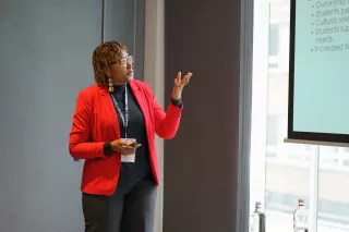 Cleopatra is in red blazer and presents confidently beside a screen displaying bullet points. Her expression is engaged, creating an informative atmosphere.