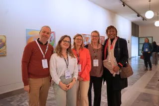 A group of five people smiling, standing side by side in an art gallery with abstract paintings on the wall. They're wearing casual business attire and conference badges, creating a friendly and professional atmosphere.