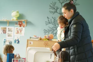 A caregiver in a puffy jacket holds a toddler in a classroom with blue walls. Kids play nearby. The atmosphere is warm and nurturing, with playful decor.