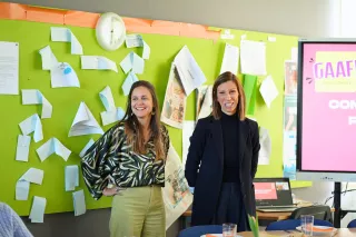 Two women stand smiling in front of a green bulletin board covered with papers. A screen beside them shows the word "GAAF." The mood is cheerful.