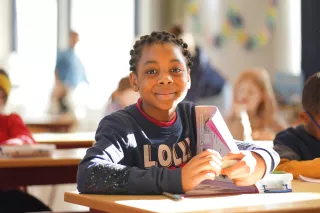 A smiling child in a classroom sits at a desk, holding a book. The sunny, bright room has other children in the background, creating a cheerful, learning atmosphere.