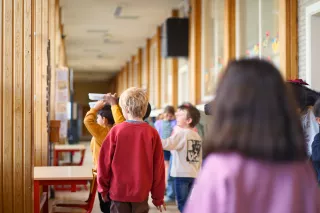 Children play happily in a school hallway with wooden walls and large windows. A child in a red sweater stands out in the lively scene.