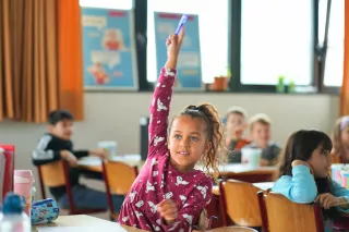 A young girl in a purple sweater eagerly raises her hand in a classroom, surrounded by classmates sitting at desks, conveying enthusiasm and engagement.
