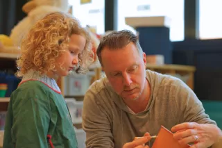 A man and child focus on painting a small pot in a classroom. The child watches intently, while the man carefully applies paint, conveying a sense of concentration and learning.