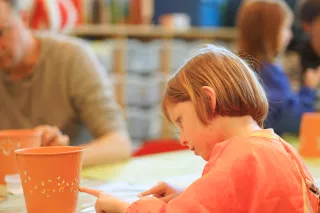A young child in a pink smock focuses intently on painting an orange pot at a table. Blurred background suggests a lively, creative classroom.