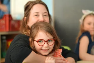A smiling young girl with red glasses is hugged by a woman, both happy. Another child is visible in the background, creating a warm, joyful tone.