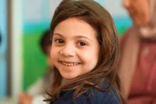 A young girl with long brown hair smiles warmly at the camera, conveying happiness. The blurred background suggests a bright, colourful classroom setting.