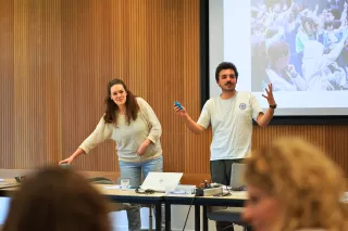 Two people are presenting in a room with wood-paneled walls. The woman, dressed casually, smiles while adjusting equipment. The man gestures animatedly with a remote. A projected image of an audience is visible. The atmosphere is engaging and interactive.
