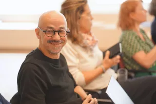 A smiling man with glasses in a black shirt sits in focus, surrounded by others in a blurred background, creating a warm and friendly atmosphere.
