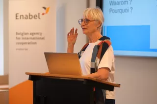 Elderly woman with glasses and short white hair giving a presentation at a podium, with "Enabel" logo and screen displaying text in the background.