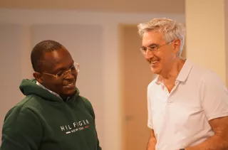 Two men are conversing and smiling indoors. One wears a green hoodie, the other a white polo shirt. The ambiance is warm and friendly.