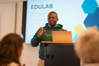 A man wearing glasses and a blazer speaks at a podium in a conference room. He gestures with his hands, conveying an engaging presentation.