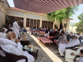 Outdoor gathering with seated men in white robes and students in uniforms. Food on tables, relaxed and attentive atmosphere under a shaded canopy.