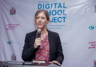 Woman speaking through a microphone, standing in front of a banner that says Digital School Project