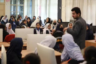 A classroom filled with students, mostly young women wearing headscarves, attentively listening to a male instructor. The atmosphere is focused and engaged.