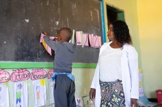 A young boy hangs a paper number on a classroom chalkboard, while teacher Emelda in a white coat watches and supports him. Colourful drawings and month labels decorate the wall below the board