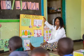 Teacher Emelda sits on the classroom floor showing a colourful choice board to a group of young learners.