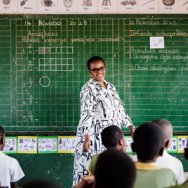 A teacher in a South African classroom stands in front of a chalkboard, while students look ahead.