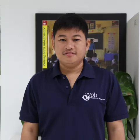 Sokhoeun wearing a navy VVOB polo shirt, standing in front of a framed picture of students in a classroom.