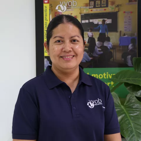 Chandaphea wearing a navy VVOB polo shirt, standing in front of a framed VVO picture of a classroom.