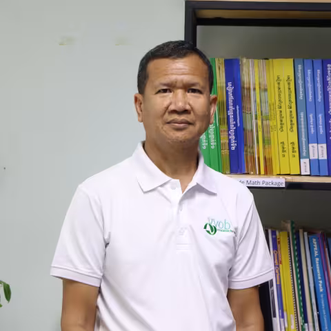 Brohs wearing a white t-shirt with the VVOB logo on it in green. He is standing in front of a bookshelf filled with blue and yellow books.
