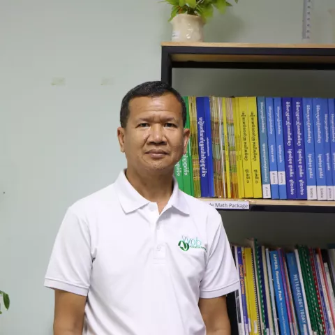 Brohs wearing a white t-shirt with the VVOB logo on it in green. He is standing in front of a bookshelf filled with blue and yellow books.