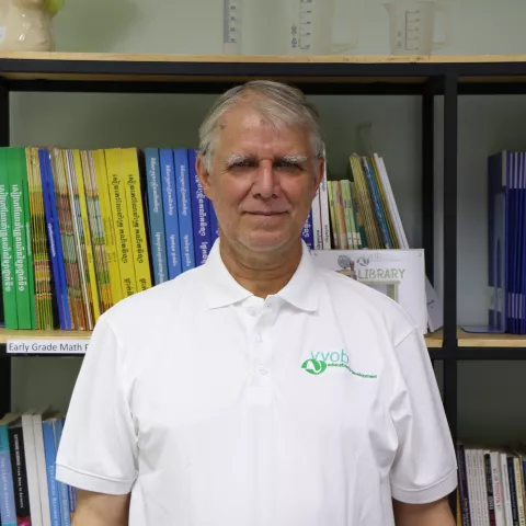 Carl wearing a white t-shirt with the VVOB logo on it in green. He is standing in front of a bookshelf filled with blue and yellow books.