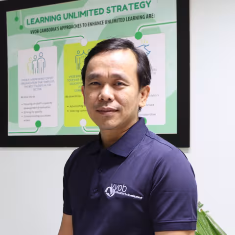 Vannarith wearing a navy VVOB polo shirt, standing in front of a framed picture with VVOB's Learning Unlimited Strategy.
