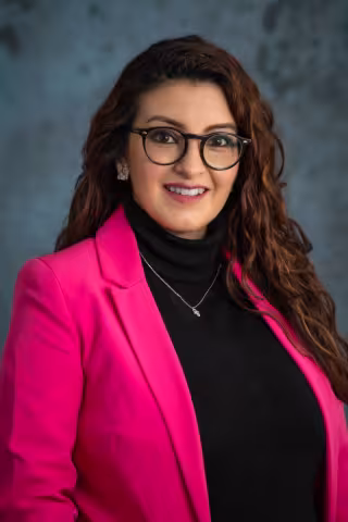 Belen smiles softly looking into the camera. She wears a pink blazer over a black shirt. The background is a mottled blue.