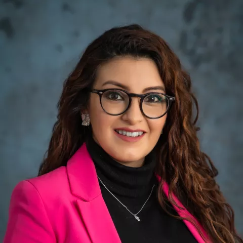 Belen smiles softly looking into the camera. She wears a pink blazer over a black shirt. The background is a mottled blue.