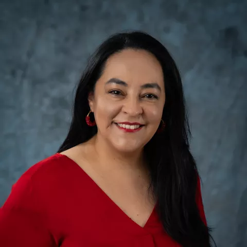 Carla smiles as the camera. She wears a red shirt. The background is a malted blue backdrop. 
