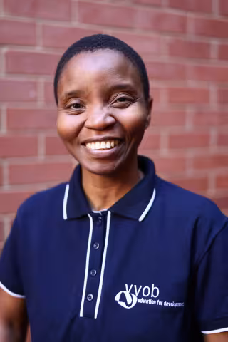 Celiwe smiling in a navy polo shirt stands against a brick wall. Her expression is warm and friendly.