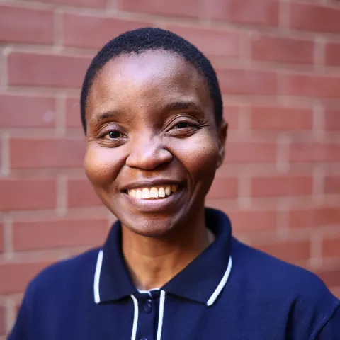 Celiwe smiling in a navy polo shirt stands against a brick wall. Her expression is warm and friendly.