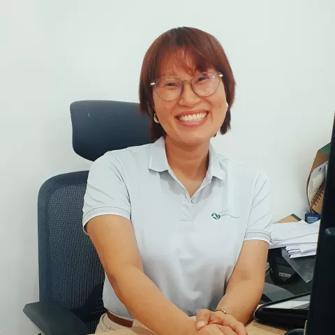 Thin smiles warmly in a white VVOB polo shirt and glasses. She is sitting on a black office chair against a while wall. 