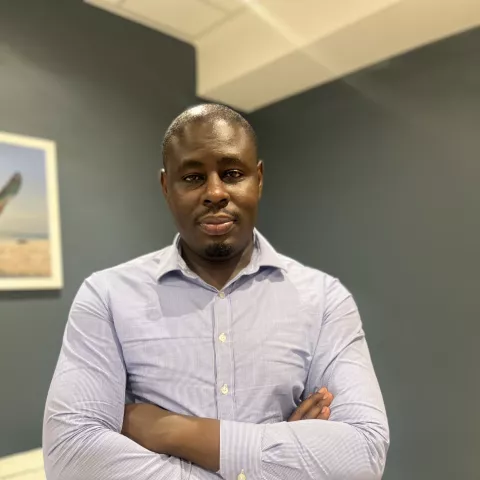 Christopher is wearing a light blue shirt and stands confidently with arms crossed in a living room. Behind him is a painting of a boat.